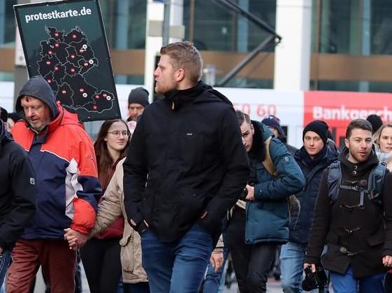 Carolin Lichtenheld und Martin Schieck auf einem Querdenken Aufmarsch in Magdeburg. Schieck hält eine Kamera in der rechten Hand. Neben Lichtenheld wird ein Plakat mit "protestkarte.de" getragen.