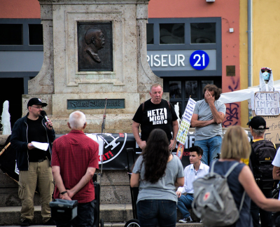 Bei einer Montagsdemo stehen auf dem Jenaer Marktplatz Teilnehmer*innen vor dem Brunnen, auf dessen Stufen Axel Robert Göhring am Mikrofon redet und Dante Riedel weiter rechts sitzt.