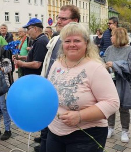 Manuela Behrend steht mit einem blauen Luftballon in der Hand auf dem Schleizer Marktplatz und schaut in die Kamera. Im Hintergrund ist u.a. Hartmut Lucas zu sehen.