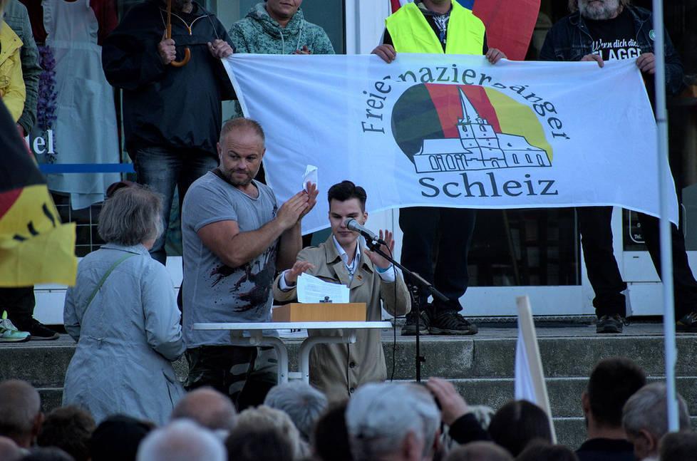 Foto von Christian Klar und Dante Riedel am Mikrofon vor der Kundgebung. Im Hintergrund eine Fahne von "Freie Spaziergänger Schleiz"