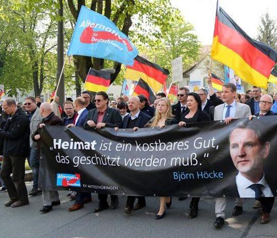 Stefan Möller, Stephan Brandner, Andreas Kalbitz, Wiebke Muhsal, Birgit Bessin, Björn Höcke und Oliver Kirchner tragen ein schwarzes Transparent auf dem der Text steht: "'Heimat ist ein kostbares Gut, das geschützt werden muss.' Björn Höcke". Daneben ist ein Porträtfoto von Höcke abgebildet.