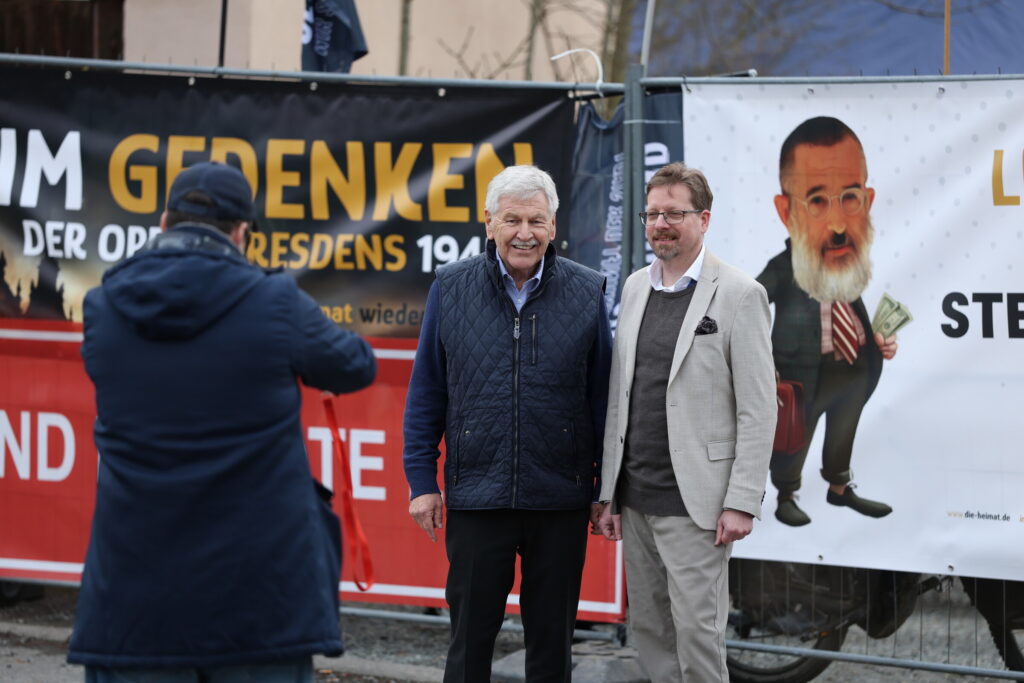 Udo Voigt und Peter Schreiber posieren vor dem Zaun vor dem neuen Parteibüro. Eine dritte Person macht ein Foto von ihnen.