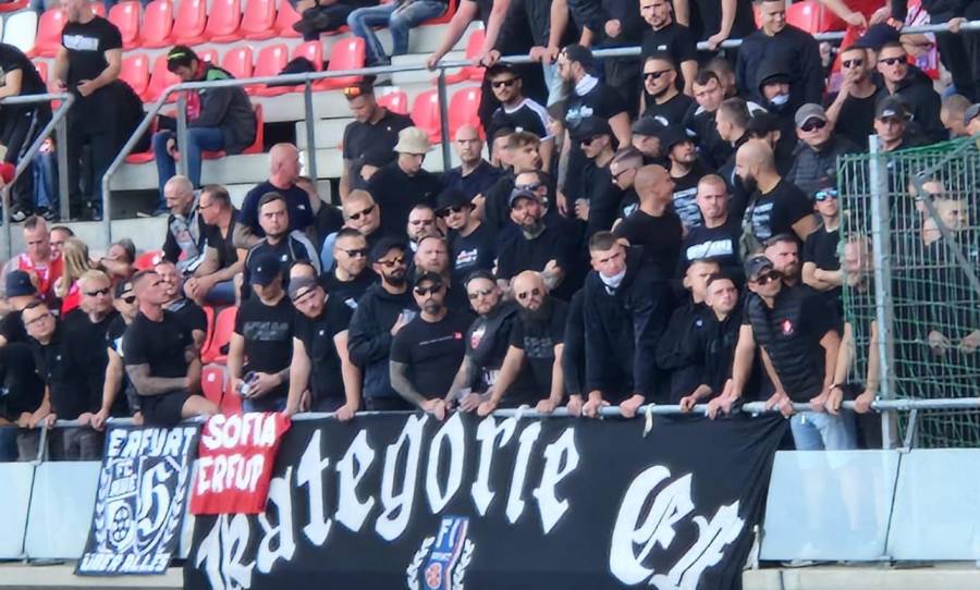 Auf der Tribüne des Steigerwaldstadions stehen ca. 30 Neonazi-Hooligans, alle in schwarz oder schwarz-weiß gekleidet. Am Geländer haben sie ein großes schwarzes Banner "Kategorie EF", ein kleines schwarzes Banner "Erfurt über alles" und eine noch kleinere rote Zaunfahne "SOFIA ERFURT" befestigt.