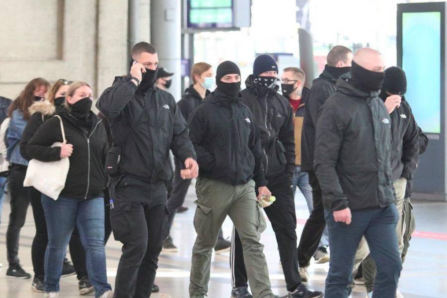Eine Gruppe schwarz gekleidert und vermummter Neonazis läuft durch den Leipziger Bahnhof. Links ist Kevin Noeske zu erkennen, der ein Telefon am Ohr hält. Mittig läuft Paul Voß, der eine Cargo-Hose in olivgrün trägt.