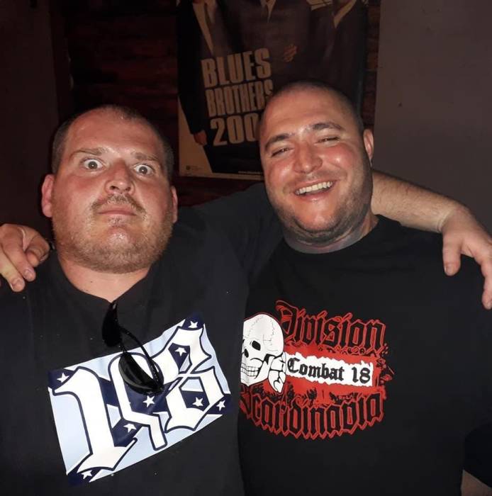 Zwei Neonazi-Skinheads blicken Arm in Arm in die Kamera. Rechts ist Orlin Pavlovzu sehen, der ein Shirt vom "Combat 18 Scandinavia" trägt, auf dem ein SS-Totenkopf zu sehen ist.