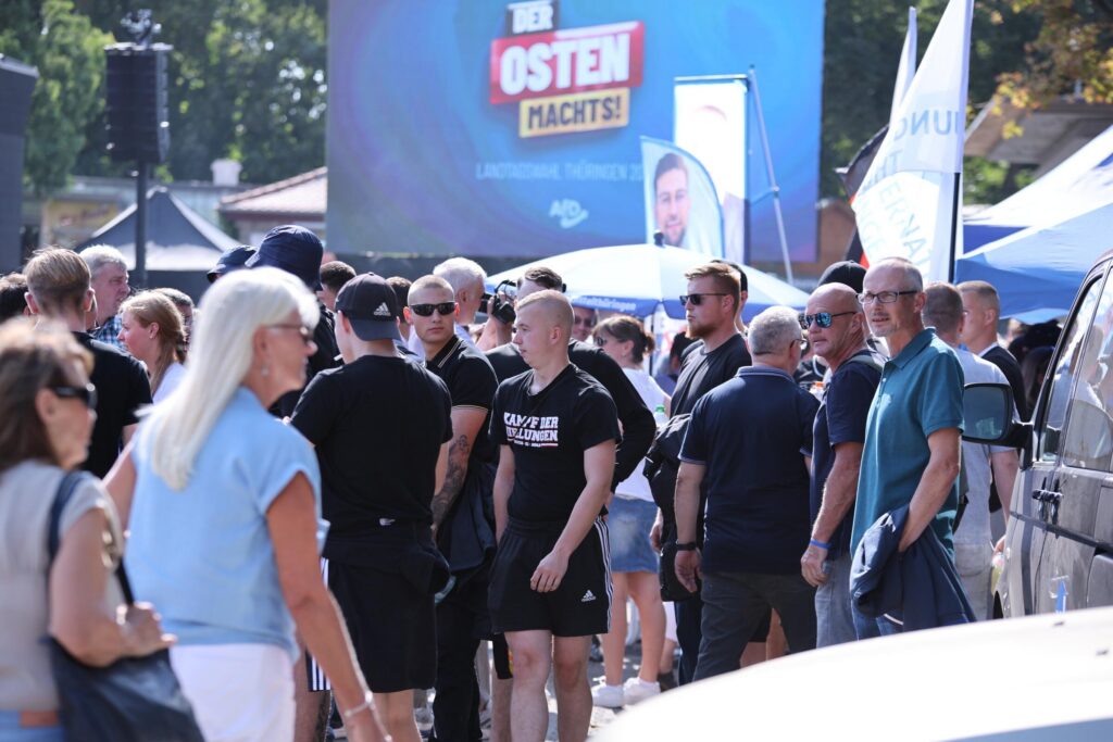 Man sieht eine Gruppe jugendlicher bis junger Männer mit kurzen Haaren auf dem Erfurter Domplatz. Im Hintergrund ist die AfD-Bühne mit dem Wahlkampfmotto "Der Osten macht's" zu sehen. Mittig sieht man Robin Klein mit Shorts und einem schwarzen Shirt mit der Aufschrift "Kampf der Nibelungen".