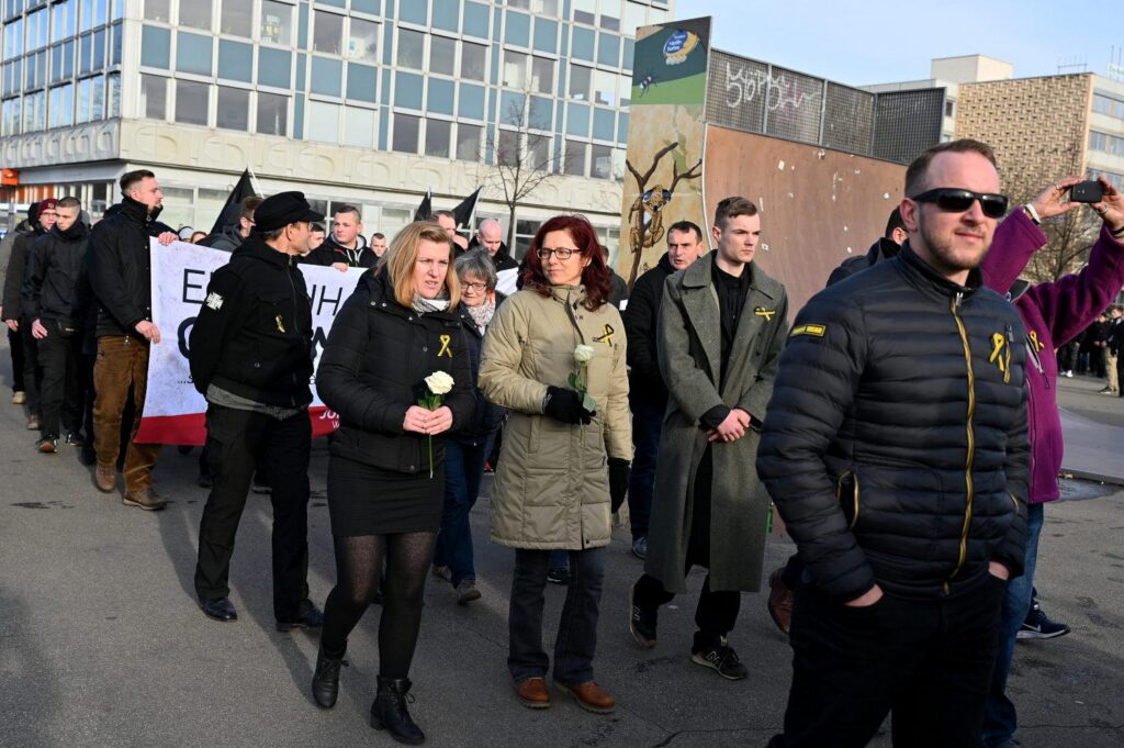 Man sieht Beatrice Fischer und eine weitere Frau mit jeweils weißen Blumen in der Hand. Rechts von ihnen läuft Etienne Klupp in einem langen Mantel. Es ist der Start des Dresdener Naziaufmarschs 2020 vor einer Halfpipe an einem Skatepark.