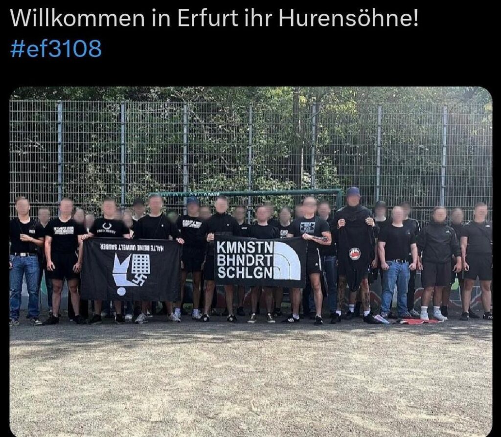 Auf einem Sportplatz stehen ca. 20 junge und in schwarz gekleidete Männer, deren Gesichter verpixelt sind. Links wird eine Anti-Nazi-Fahne verkehrt herum gehalten, mittig ein Banner mit dem im Stil von "The North Face" gehaltenen Schriftzug "KMMNSTN BHNDRT SCHLGN". Darüber wird kommentiert: "Willkommen in Erfurt ihr Hurensöhne #3108"
