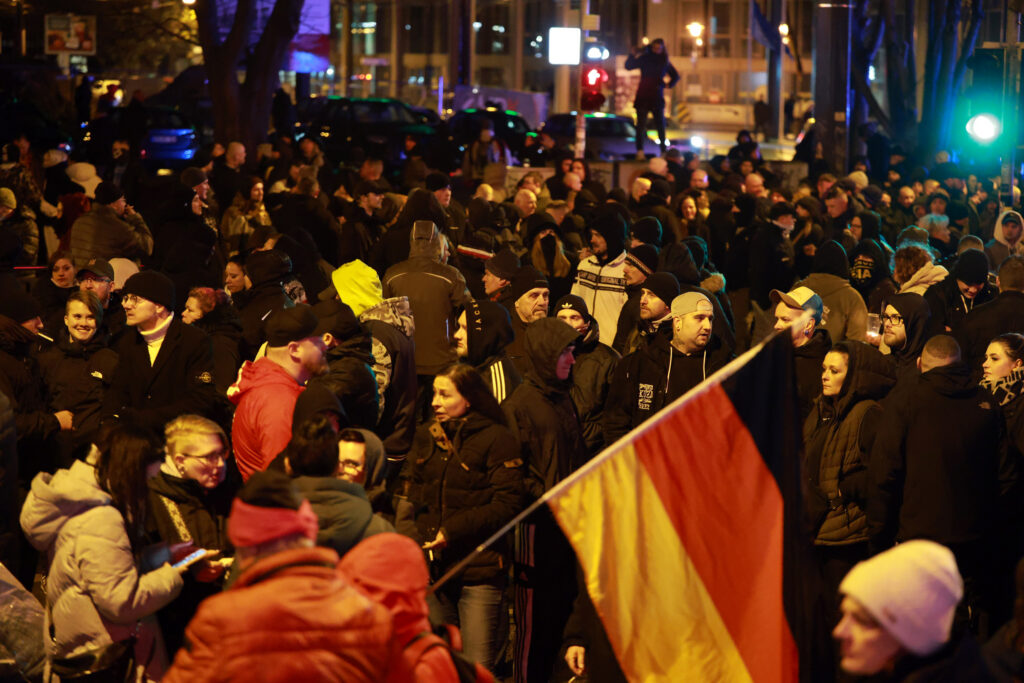 Man sieht Hunderte, oft junge Neonazis im Dunkeln auf dem Magdeburger Hasselbachplatz. Vorne wird eine Deutschlandfahne gehalten. Am linken Bildrand sieht man Caroline Hein lachend mit einer schwarzen Jacke.