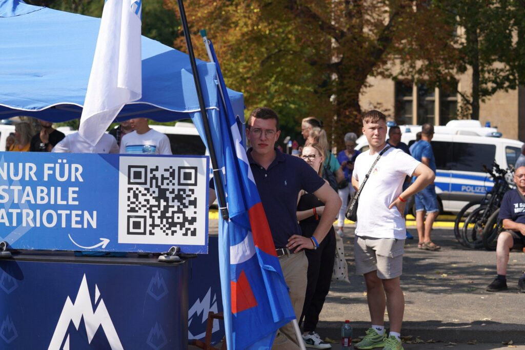 Foto von Julian und Haily-Marie Kitz sowie Timm Westermann (1.v.l.) beim Wahlkampfabschluss der AfD in Erfurt am 31.08.2024. Julian und Kitz stehen hinter einem blauen Infostand. Westermann lehnt an diesem, vor ihm stehen blaue AfD-Fahnen. Rechts im Bild ist eingroßer QR-Code zu sehen, der mit „Nur für stabile Patrioten“ beschriftet ist. Unten am Infostand ist das damalige Logo der Jungen Alternative zu sehen.