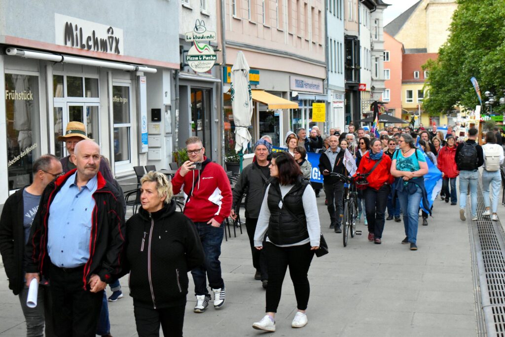 Foto eines PandemieleugnerInnenaufmarsch, der durch die Jenaer Johannisstraße läuft. Im Vordergrund läuft Volker Wagenhaus Hand in Hand mit einer anderen Person. Dahinter laufen einige weitere Personen, die ein Transparent tragen, dessen Beschriftung nicht lesbar ist.