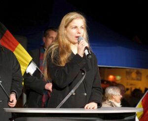 Foto von Wiebke Muhsal an einem Redner*innenpult. Sie hält ein Mikrofon in der Hand. Im Hintergrund steht eine Person mit einer schwarz-rot-goldenen Fahne.