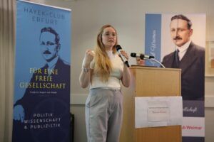 Foto von Michaela Meißner, die an einem Rednerpult steht und in ein Mikrofon spricht. Sie trägt ein weißes Shirt, eine Brille und eine graue Hose. Im Hintergrund ist ein blauer Aufsteller des Hayek-Clubs Erfurt zu sehen. Darauf steht: Hayek-Club Erfurt. Für eine freie Gesellschaft. Friedrich August von Hayek. Politik, Wissenschaft & Publizistik.