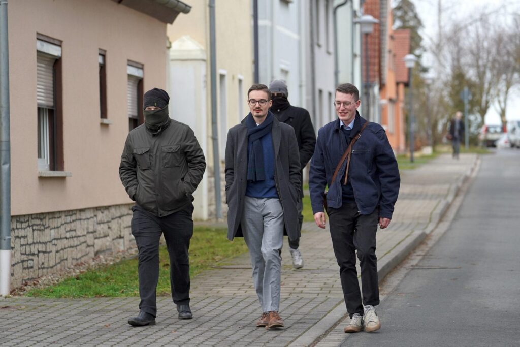 Foto von Friedrich Hunger und Timm-Albert Westermann bei den Studientagen in Schnellroda am 25.01.2025. Hunger läuft in der Mitte und trägt Brille, ein blaues Oberteil, eine graue Hose und einen grauen Mantel. Rechts von ihm ist Westermann zu sehen, hinter und links von Hunger laufen zwei vermummte, dunkel gekleidete Personen.