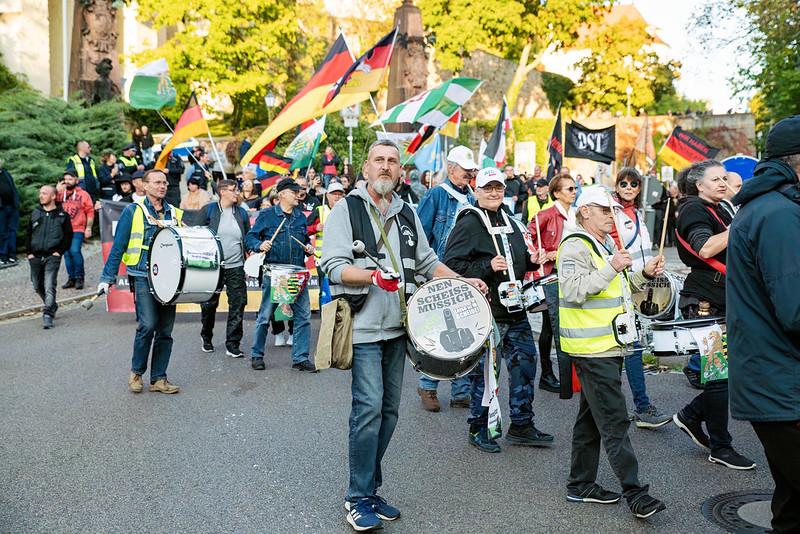 Foto von Angela Lehmann beim Neonazi-Aufmarsch von „Freies Thüringen“ und „Freies Sachsen“ in Altenburg. Lehmann trägt eine schwarze Jacke, eine weiße AfD-Kappe und hat eine Trommel vor sich. Um sie herum sind weitere TrommelspielerInnen. Links neben ihr läuft ein Mann, auf dessen Trommel „Nen Scheiss muss ich“ steht. Andere Teilnehmende haben Deutschlandflaggen, Sachsenflaggen und eine Fahne mit dem Kürzel „DST“ dabei.