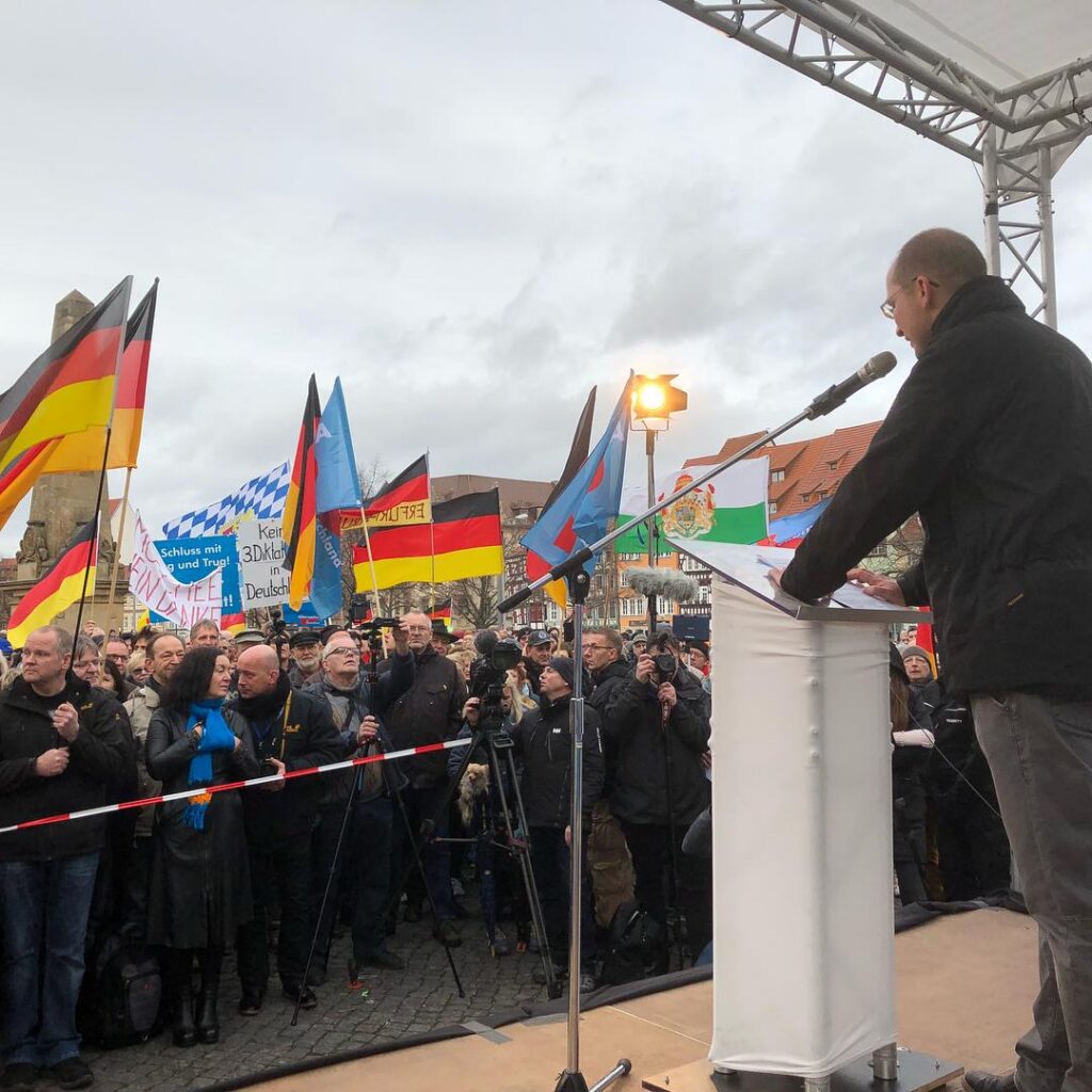 Foto von Dirk Zabel bei einer AfD-Kundgebung in Erfurt am 28.01.2018. Zu sehen ist eine Bühne auf der Stefan Möller steht. Davor stehen die Teilnehmenden hinter einem Absperrband. Als dritter von rechts steht Zabel in der ersten Reihe, gekleidet in einer schwarzen Jacke. Im Hintergund wehen Deutschland-, Bayern-, Sachsen- und AfD-Flaggen. Auf einem Schild steht „Keine 3.Diktatur in Deutschland“ sowie „Schluss mit Lug und Trug!