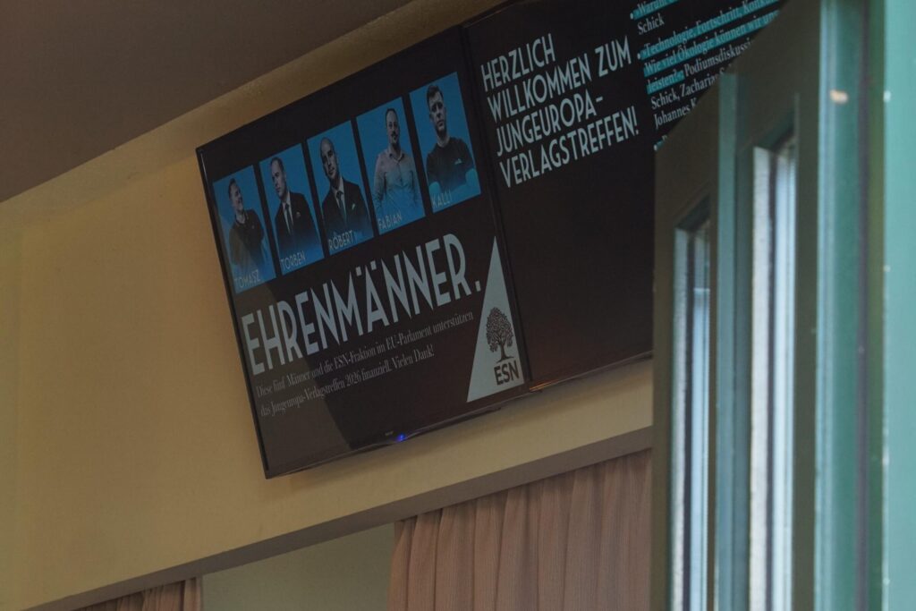 An Wand angebrachter Fernseher, der zeigt: Fünf Fotos mit den Namen Tomasz, Torben, Robert, Fabian und Kalli. Darunter in Groß "Ehrenmänner." Darunter: "Diese fünf Männer und die ESN-Fraktion im EU-Parlament unterstützen das Jungeuropa-Verlagstreffen 2026 finanziell. Vielen Dank!" In der rechten unteren Ecke das Logo der ESN.