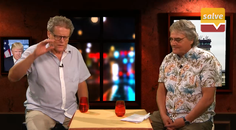Man sieht Norbert Dotterweich und Klaus-Dieter Böhm-Schneider in kurzärmligen Hemden im Salve TV Studio im Gespräch. Die beiden stehen sich an einem kleinen Holztisch gegenüber, auf diesem sind zwei orangene Gläser sowie ein Stapel Papiere platziert, die Böhm-Schneider vor sich abgelegt hat. Er trägt ein buntes Hawaihemd und eine schwarze Uhr am linken Handgelenk, sein Blick wandert nach unten zu seinen Notizzetteln. Dotterweich ihm gegenüber trägt ein weites hellblaues Hemd, sein rechter Arm ist im Redeschwall leicht erhoben. Links hinter ihm ist auf einem Bildschirm oder Bild Donald Trump vor einer US-Fahne zu erkennen.