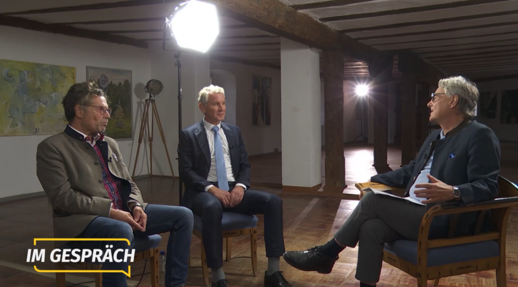 eine Bildmontage. Links: Zu sehen ist eine große Menschenmenge. Im Hintergrund sind verschiedene Fahnen zu erkennen: zwei Deutchlandfahnen, mehrere Thüringenfahnen sowie eine Fahne mit dem Loge von „Freies Thüringen“. Außerdem werden zwei Schilder hochgehalten, das Hintere ist zu weit in der Ferne um die Schriftzeichen darauf zu erkennen, auf dem Vorderen steht in schwarz und orange (den Fahnen von „Die Heimat“) „Frieden mit Russland“. Vorne links steht eine lächelde Marion Schneider, in den Händen ein wohl die Bühne vor ihr filmendes Handy. Rechts: Die drei Männer sitzen jeweils auf Holzstühlen mit blauem Polster. Rechts Dieter Böhm-Schneider in einem Sessel. Seine Beine sind überschlagen, auf diesen liegen mehrere Blätter Papier. Böhm-Schneiders Blick ist auf den mittig sitzenden Björn Höcke gerichtet, dieser trägt als einziger eine überdimensionierte blaue Krawatte. Hinter Höcke steht ein großer LED-Strahler, der sich im Hintergrund spiegelt. Am linken Bildrand sitzt Dieter Dehm in Trachtenjacke.
