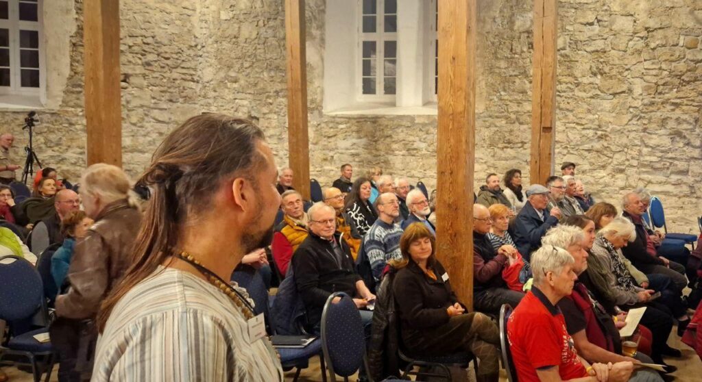 Man sieht den Kongresssaal aus dem Jahr 2025. Die Teilnehmenden sitzen in dichten Stuhlreihen hintereinander. Rechts vorne sitzt Winfried Merkel in einem roten Shirt. Im Vordergrund des Bildes steht ein Mann mit langen Haaren und einem grauen Oberteil.