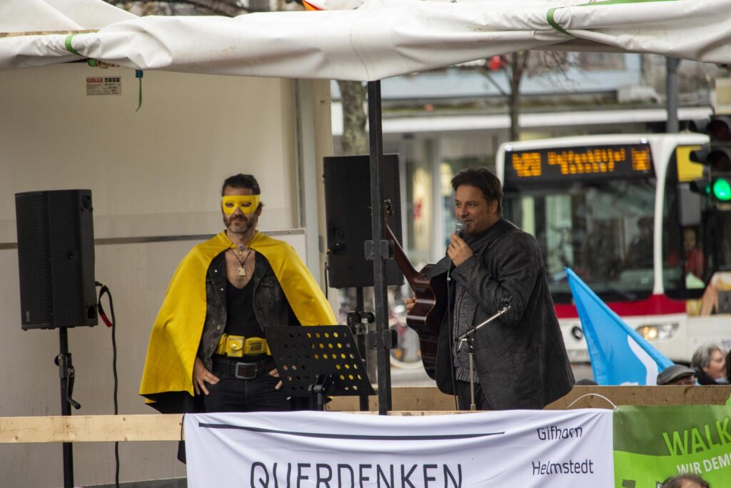 Krengel steht auf einer Bühne mit weißem Dach und hält eine Gitarre sowie ein Mikrofon, in das er spricht. Neben ihm steht ein Mann in gelber Verkleidung. An der Bühne hängt ein Transparent mit der Aufschrift: "Querdenken".