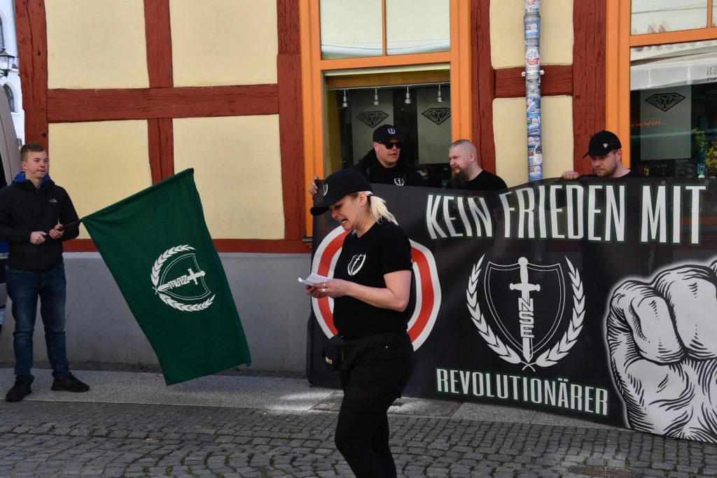 Robin Klein steht mit grüner Fahne der „Neue Stärke Partei“ und Schlauchschal am Hals in einer Kundgebung. Mittig ist Sabrina Töpfer bei einer Rede am Mikrofon zu sehen. Im Hintergrund stehen mehrere Neonazis hinter einem Banner.