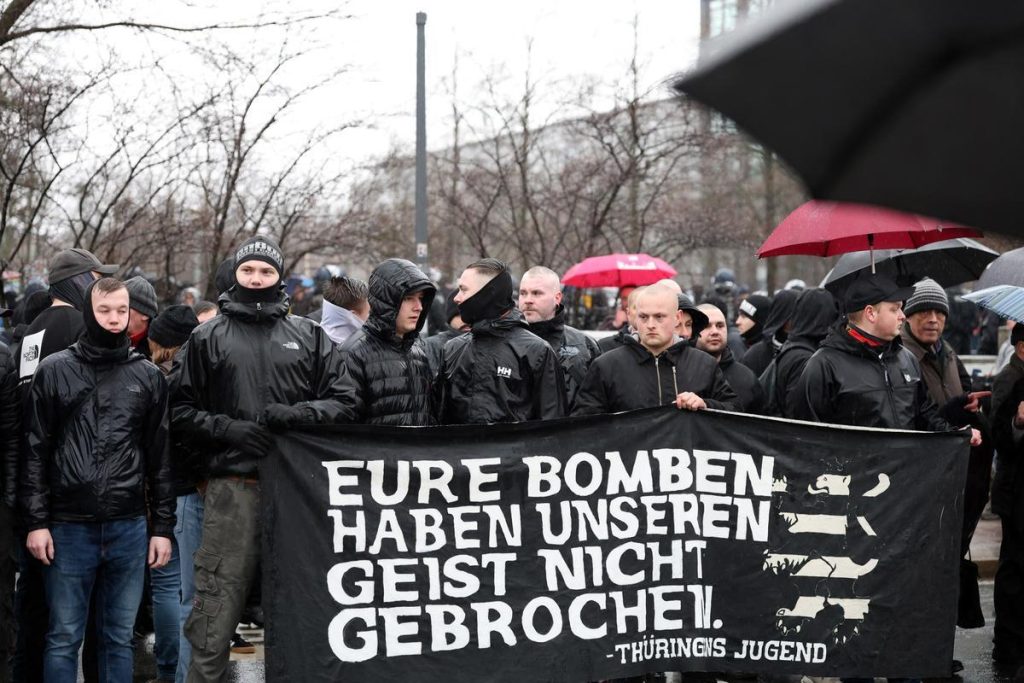 Eine Gruppe junger Männer in schwarzer Outdoor-Kleidung steht nassgeregnet auf dem Dresdener Naziaufmarsch. Sie halten ein schwarz-weißes Banner mit dem Spruch: „Eure Bomben haben unseren Geist nicht gebrochen“. Robin Klein steht links und hat einen Schlauchschal halb übers Kinn gezogen.