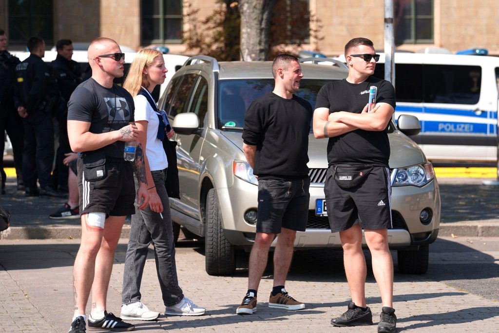 Man sieht vier Personen auf dem Erfurter Domplatz. Links steht Eric Krempler mit Glatze und Sonnenbrille, daneben seine damalige Freundin Ida Wolf mit offenen blonden Haaren und weißem Shirt. Daneben steht ein Mann in schwarzer Kleidung und rechts Danilo Martin mit kurzer Scheitelfrisur, Shirt von „Label 23“, Sonnenbrille und verschränkten Armen. Bildunterschrift: „Danilo Martin (1·v.r.) mit Eric Krempler (1.v.l.) und Ida Wolf (2.v.l.) auf dem AfD-Wahlkampfabschluss in Erfurt am 31.8.2024 (Foto: Recherche Nord)“