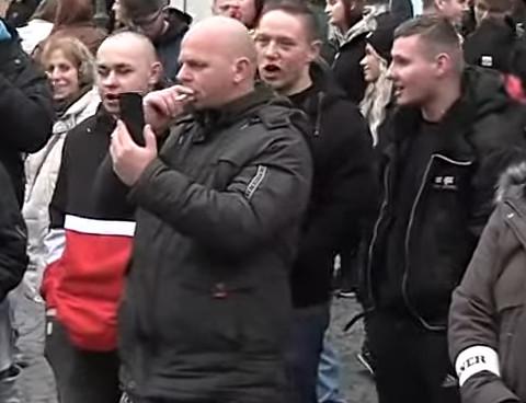 Screenshot aus einem Youtube-Stream aus Greiz, auf dem vorne mit Glatze Andy Fahl zu sehen ist, dahinter Ian Bütow mit Glatze und schwarz-weiß roter Jacke und daneben Kevin Pöllinger und Benny Franke, beide in schwarz. Alle haben beim Parolen rufen die Münder offen.