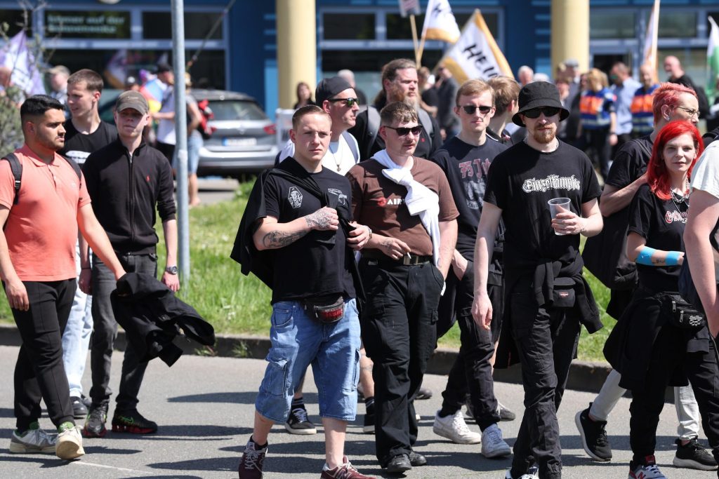 Foto vom Naziaufmarsch in Gera am 1. Mai 2025, auf dem Pöllinge rin schwarzem Shirt, kurzer Hose und Scheitelfrisur im Vordergrund zu sehen ist. Um ihn herum sind weitere Nazis in Szene-Shirts zu sehen. Im Hintergrund tragen Teilnehmer Flaggen von „Die Heimat“.