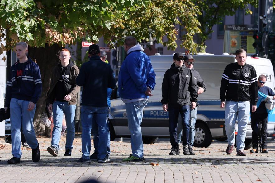 Arthur Jäcksch ist ganz rechts in Jeans und schwarzer Trainingsjacke mit weißen Querstreifen in Erfurt zu sehen. Neben ihm läuft THomas Holzinger mit Sonnenbrille und schwarzer Jacke. Ganz links ist Ben in Trainingsjacke und blondem Seitenscheitel zu sehen.