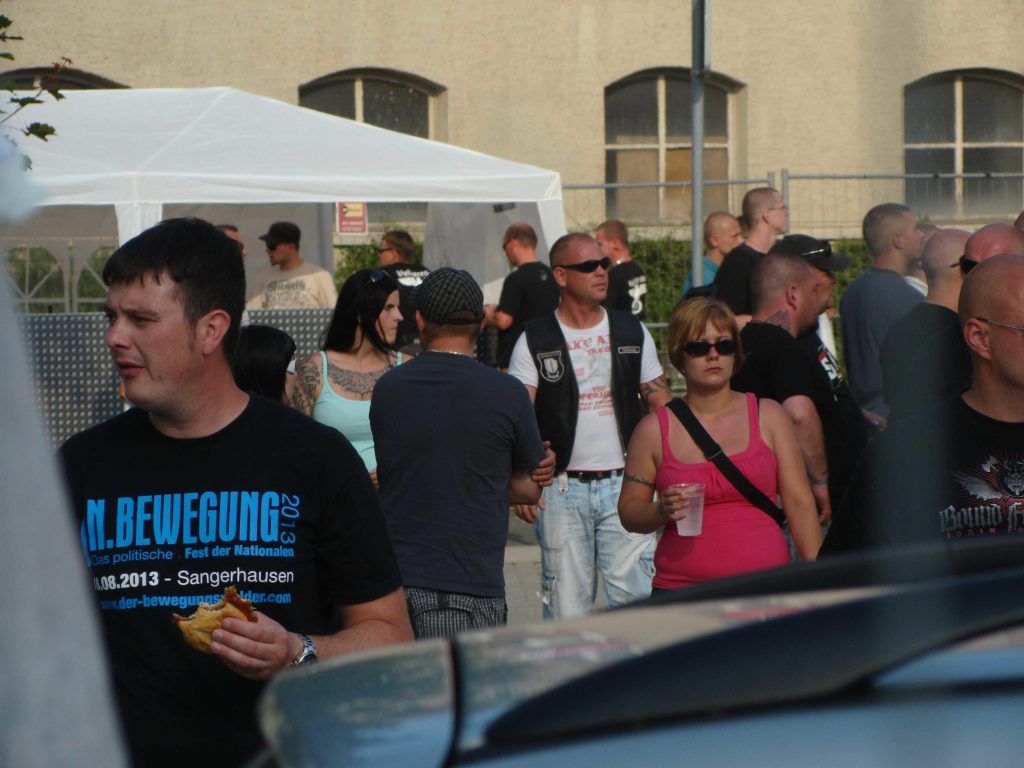Heidi Lorber ist in einem türkisen Top und offenen schwarzen Haaren neben Ronny Linke zu sehen, der über einem weißen Shirt eine Lederkutte trägt. Im Hintergrund ist ein weißer Pavillon zu sehen, im Vordergrund Patrick Weber mit einem Shirt seines Festivals „In.Bewegung“