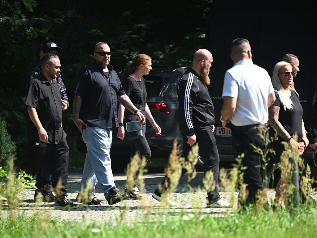 Stefan Rose ist mit längerem Vollbart, schwarzer Hose und schwarzer Adidas-Trainingsjacke zwischen weiteren schwarz gekleideten Neonazis in Beeskow zu sehen. Im Vordergrund ist in weißem Hemd und mit von der Kamera abgewendetem Blick Thorsten Heise zu sehen. Bildunterschrift: „(v.l.) Christoph Wolter (m. Cap, verdeckt), Christoph Walther („12 Golden Years“), Robert Hoyer, Stefan Rose (Trainingsjacke), Neonazi-Funktionär Thorsten Heise (in weiß), Nadine Heise und Andre Groth (1.v.r.) und am 18.8.2023 in Beeskow (Recherche Nord)“